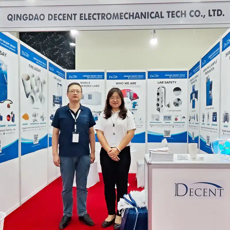 Two Qingdao Decent Group representatives, a man and a woman, standing in front of their DECENT branded booth at Minexpo Tanzania 2025, showcasing mineral assay equipment and mobile laboratory solutions.
